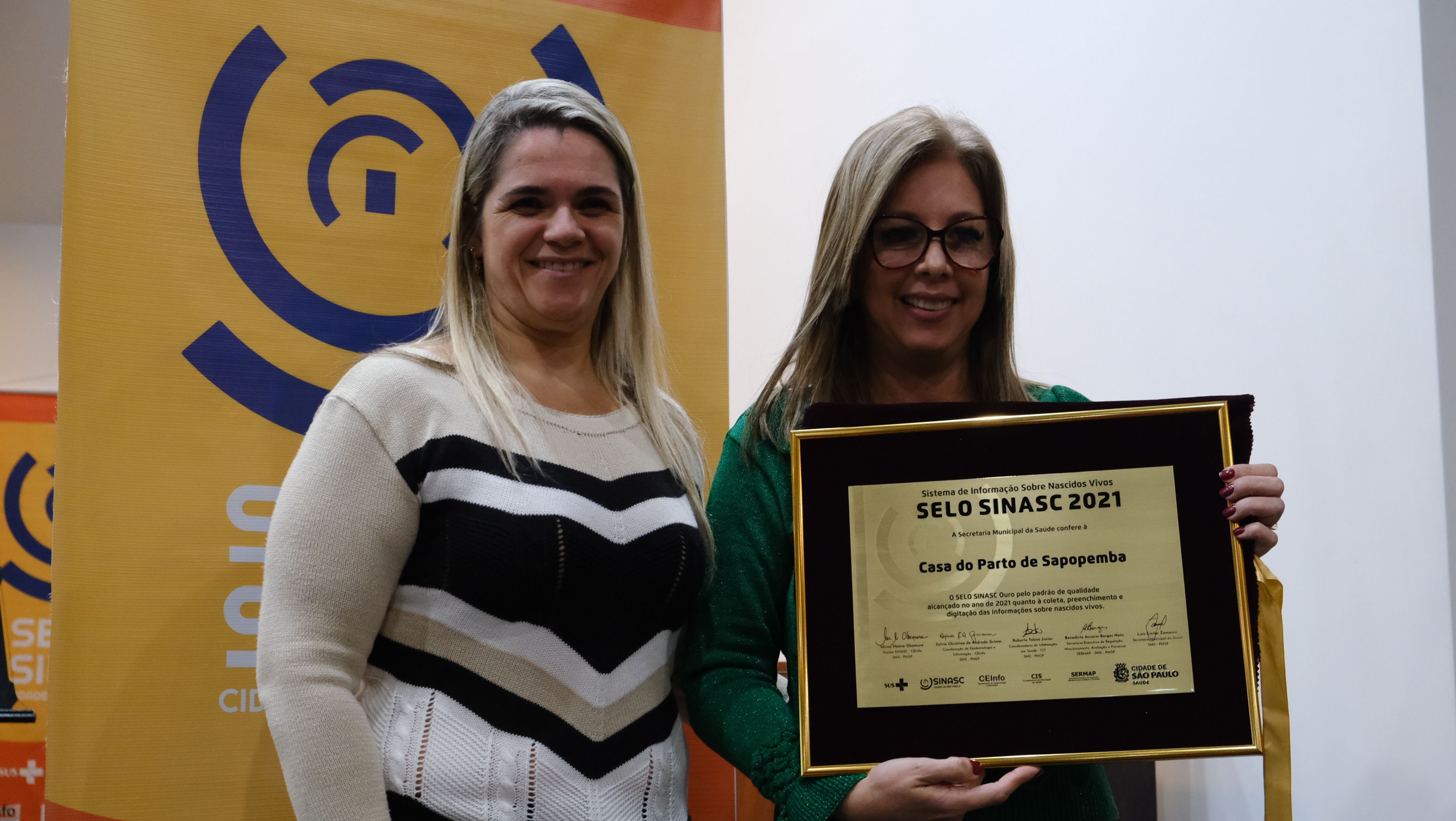 Na imagem há duas mulheres segurando uma placa de ouro. As duas estão em pé e são loiras, uma delas usa óculos e roupa verde e a outra usa roupa em tom bege com listra preta. As duas estão sorrindo.